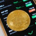 A close-up of a Bitcoin coin placed on a mobile device displaying stock market trading data.
