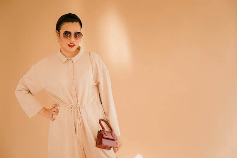 Woman posing fashionably in beige attire with sunglasses and handbag against a minimalist backdrop.